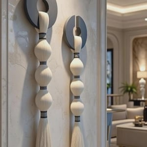 May include: Two wall-mounted decorative pieces. Each features a dark gray circular element with a vertical slit, from which a white, tassel-like design hangs. The design includes three stacked, spherical elements and a long tassel.