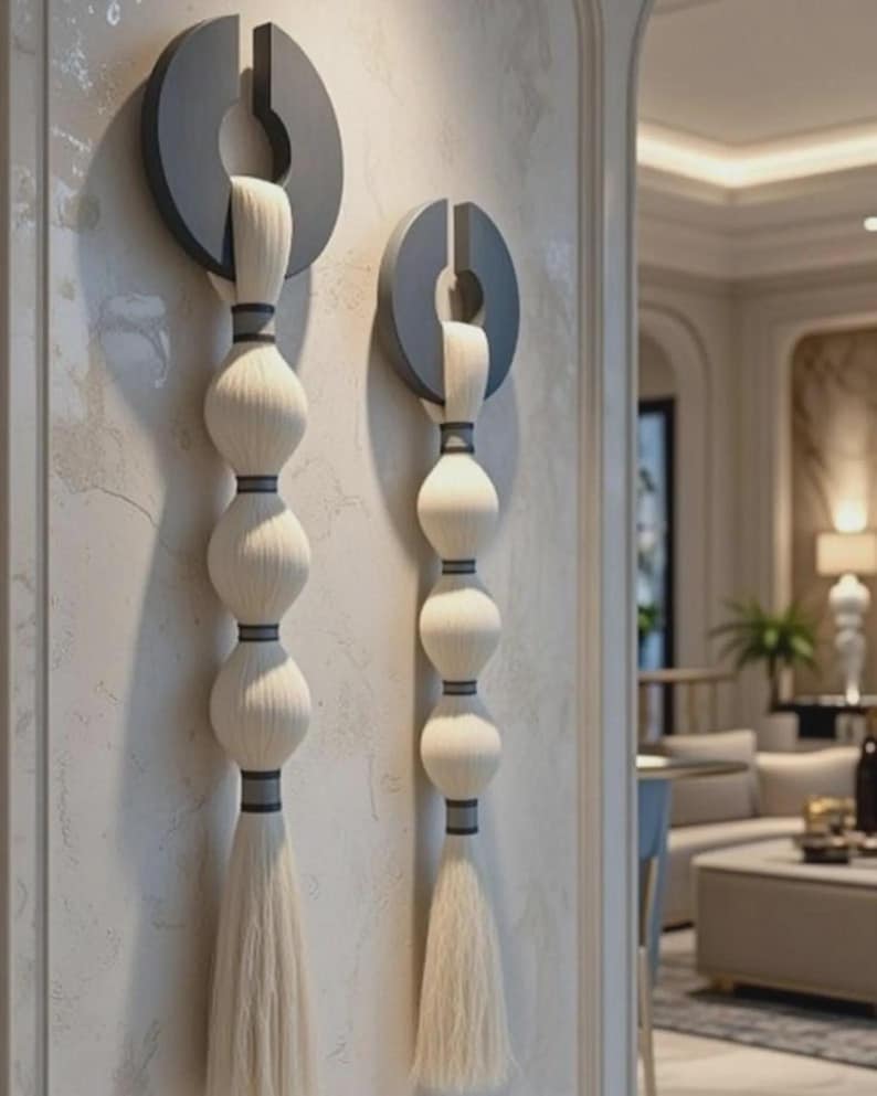 May include: Two wall-mounted decorative pieces. Each features a dark gray circular element with a vertical slit, from which a white, tassel-like design hangs. The design includes three stacked, spherical elements and a long tassel.