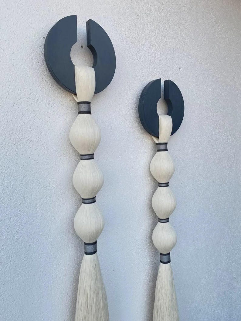 May include: Two abstract wall hangings. Each features a dark gray, semi-circular top with a vertical slit, connected to a series of white, bulbous shapes. The shapes are separated by dark gray bands, and end in a long, white tassel. Modern home decor.