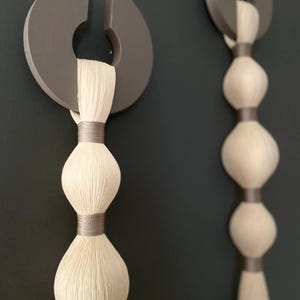 Puede incluir: Dos colgantes de pared abstractos. Cada uno presenta un elemento de madera gris semicircular con un recorte, del que cuelga un diseño de cuentas texturizado de color crema. El diseño está acentuado con una banda gris. El fondo es gris oscuro.