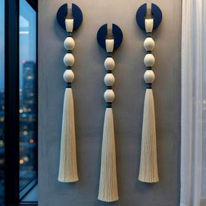 May include: Three wall hangings with a modern design. Each features a navy blue semi-circle at the top, connected to a series of white spheres and a long, cream-colored tassel. The hangings are mounted on a gray wall.
