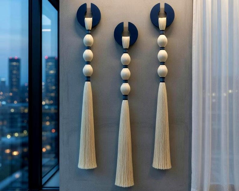 May include: Three wall hangings with a modern design. Each features a navy blue semi-circle at the top, connected to a series of white spheres and a long, cream-colored tassel. The hangings are mounted on a gray wall.