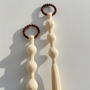 Könnte beinhalten: Zwei dekorative Wandbehänge. Jedes hat einen Holzperlenring oben, verbunden mit einer Reihe von bauchigen Formen in Creme. Einer hat eine lange Quaste unten. Der andere ist kürzer. Maße in Zentimetern.