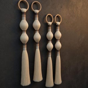 Puede incluir: Cuatro juegos de borlas decorativas con aros de madera. Cada borla presenta una serie de formas blancas y bulbosas separadas por bandas grises y marrones, que conducen a un largo fleco. Las borlas se muestran sobre un fondo gris oscuro.