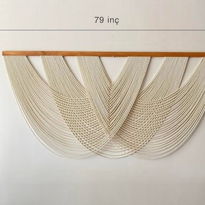 May include: A white macrame wall hanging with a wooden dowel at the top. The hanging is 79 inches wide and 40 inches tall.