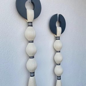 May include: Two abstract wall hangings. Each features a dark gray, semi-circular top with a vertical slit, connected to a series of white, bulbous shapes. The shapes are separated by dark gray bands, and end in a long, white tassel. Modern home decor.