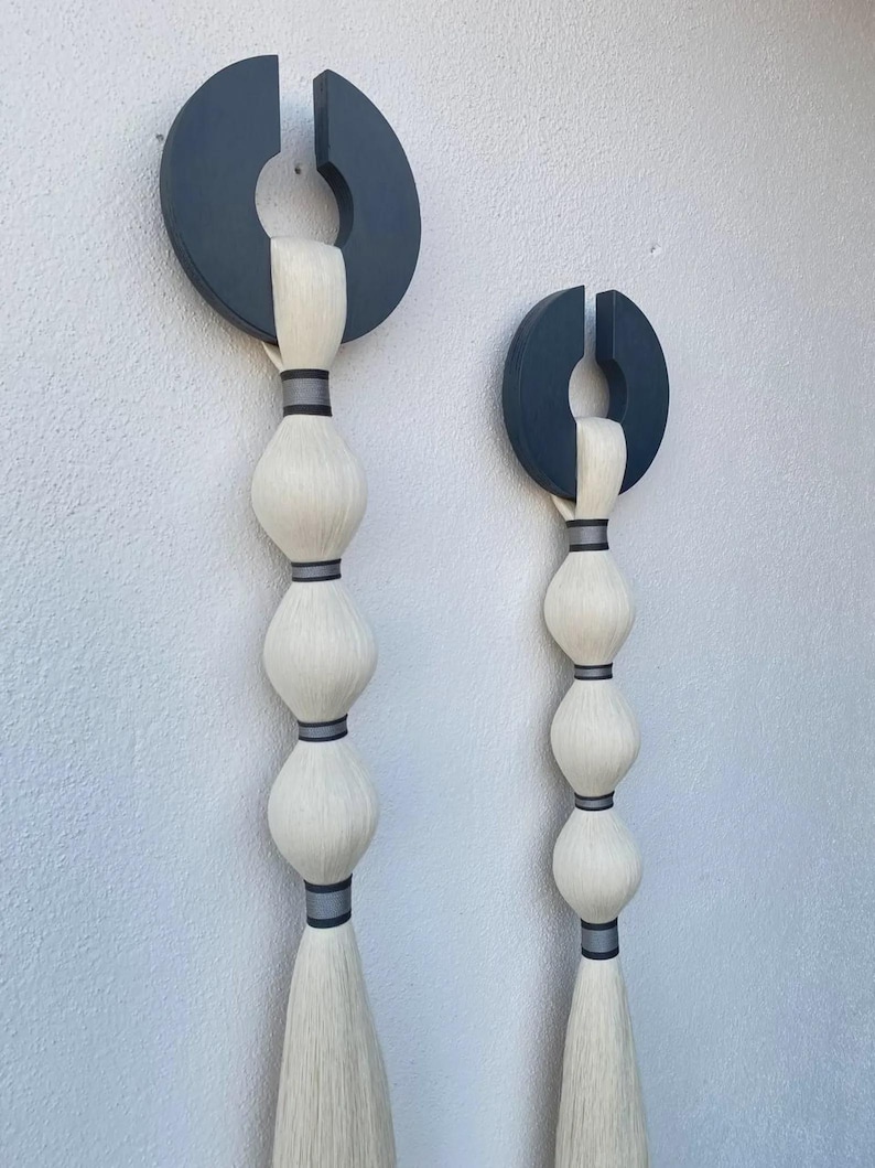 May include: Two abstract wall hangings. Each features a dark gray, semi-circular top with a vertical slit, connected to a series of white, bulbous shapes. The shapes are separated by dark gray bands, and end in a long, white tassel. Modern home decor.