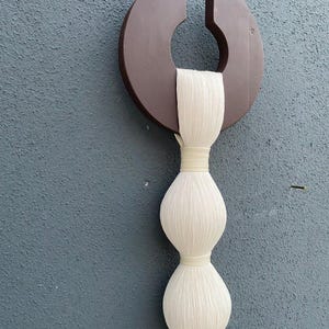 Op de afbeelding: Een wandhanger gemaakt van wit garen met een bruine houten cirkel aan de bovenkant. Het garen is geweven in een reeks van drie ronde vormen.