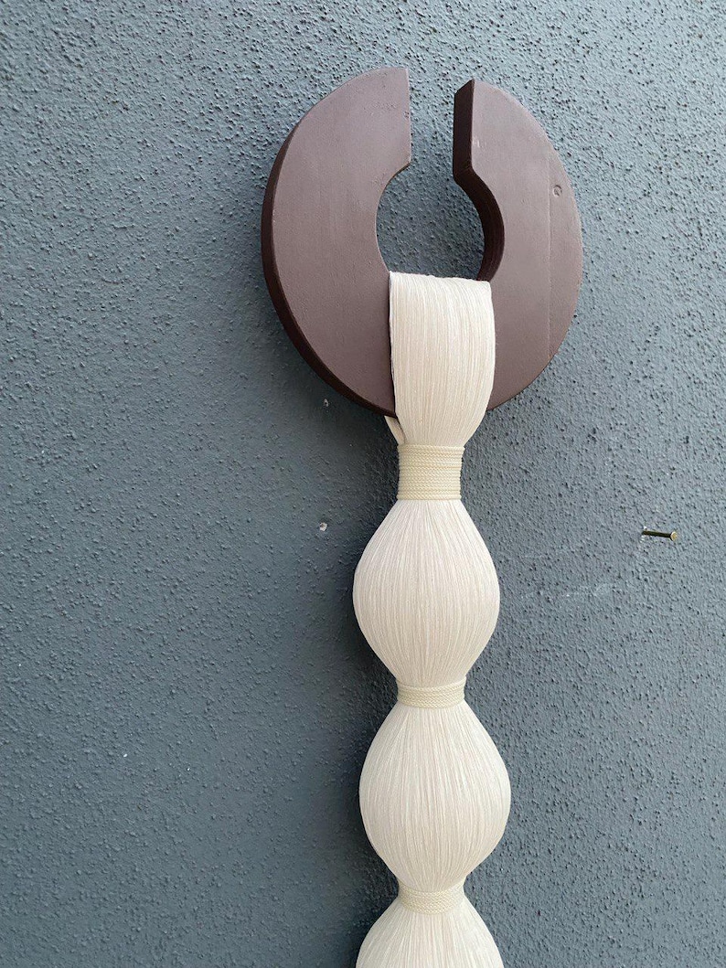 Op de afbeelding: Een wandhanger gemaakt van wit garen met een bruine houten cirkel aan de bovenkant. Het garen is geweven in een reeks van drie ronde vormen.