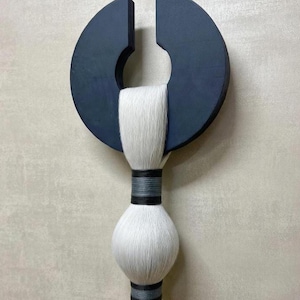 May include: A modern wall hanging featuring a dark blue, semi-circular wooden element with a central cutout. A white, textured, cylindrical element is suspended below, accented with dark gray and black bands. The piece has a minimalist, abstract design.