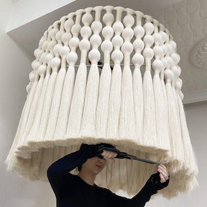Puede incluir: Una lámpara grande, de color blanco roto, hecha a mano, con un diseño único. La pantalla presenta una serie de cuentas esféricas blancas en la parte superior, con largas y fluidas borlas colgando. La forma general es una cúpula redondeada.