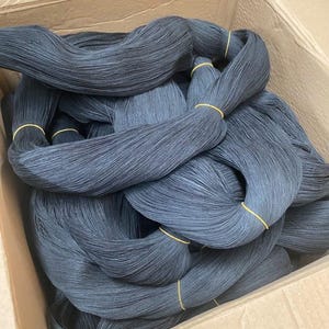 Op de afbeelding: Een kartonnen doos gevuld met bundels zwart garen. Elke bundel is vastgezet met een gele elastiek. Het garen is strak opgerold en geschikt voor diverse handwerkprojecten. De doos is open.