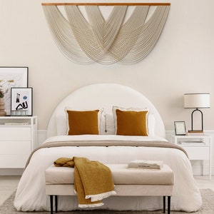May include: A white bed with a curved headboard and a white and grey patterned bedspread. Two mustard yellow throw pillows are on the bed. A mustard yellow throw blanket is draped over a white bench at the foot of the bed. A white bedside table with a black lamp is on the right side of the bed. A large white macrame wall hanging is above the bed.