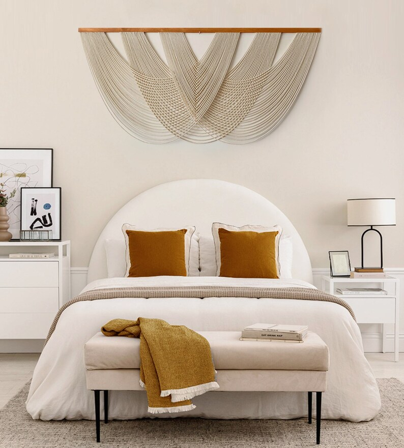 May include: A white bed with a curved headboard and a white and grey patterned bedspread. Two mustard yellow throw pillows are on the bed. A mustard yellow throw blanket is draped over a white bench at the foot of the bed. A white bedside table with a black lamp is on the right side of the bed. A large white macrame wall hanging is above the bed.