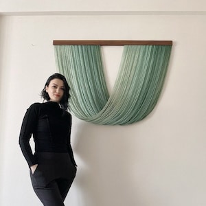 May include: A woman stands in front of a wall hanging made of green yarn. The yarn is woven into a U-shape and hangs from a wooden dowel.