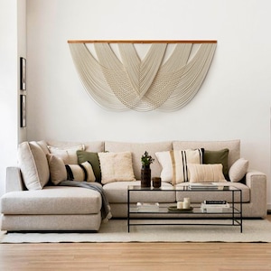May include: A beige sectional sofa with a variety of throw pillows, including green, black, and white patterns. A glass coffee table with a black metal frame sits in front of the sofa. A large macrame wall hanging with a wooden dowel hangs above the sofa.