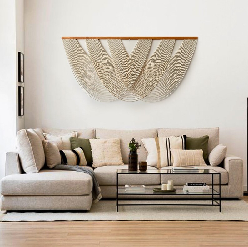 May include: A beige sectional sofa with a variety of throw pillows, including green, black, and white patterns. A glass coffee table with a black metal frame sits in front of the sofa. A large macrame wall hanging with a wooden dowel hangs above the sofa.