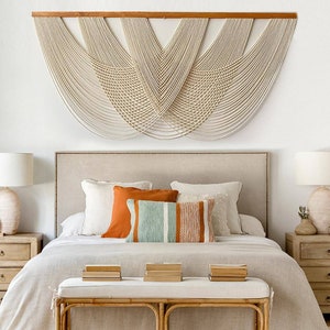May include: A bedroom with a white wall and a large macrame wall hanging above a bed with a beige headboard. The bed is made with white bedding and has a few pillows in shades of orange, white, and blue. There are two bedside tables with lamps on either side of the bed.