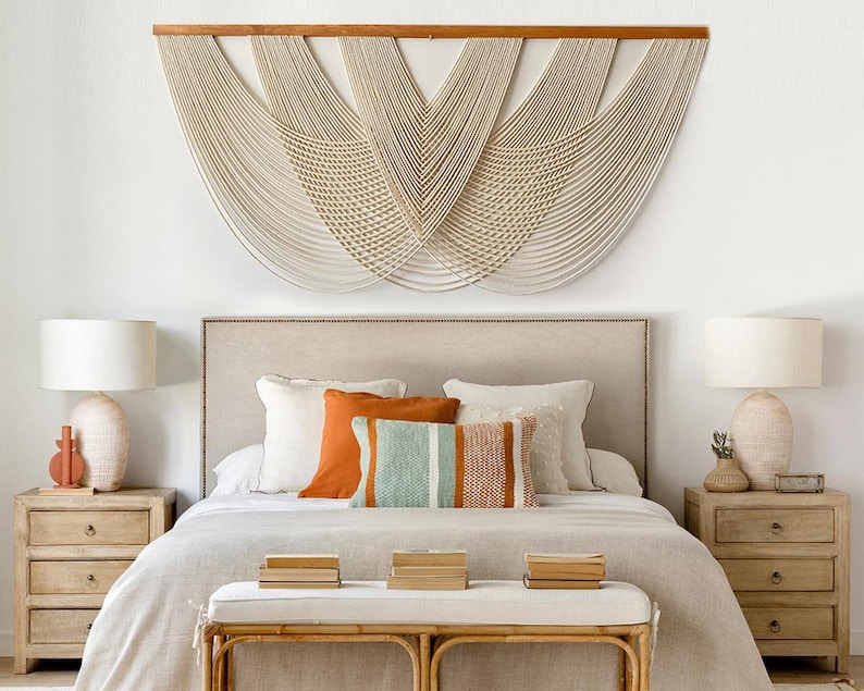 May include: A bedroom with a white wall and a large macrame wall hanging above a bed with a beige headboard. The bed is made with white bedding and has a few pillows in shades of orange, white, and blue. There are two bedside tables with lamps on either side of the bed.