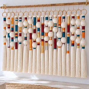 Puede incluir: Un tapiz de pared que presenta una serie de hilos verticales suspendidos de una barra de madera mediante anillos de madera. Cada hilo consta de esferas de hilo color crema, cintas de satén metálicas y de colores rojo, naranja, azul y plata, y borlas largas.