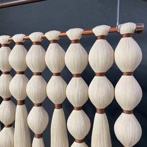 Op de afbeelding: Een witte macram&eacute; wandhanger met bruine lint accenten. De hanger is gemaakt van geknoopt garen en heeft een koperen pijp aan de bovenkant.