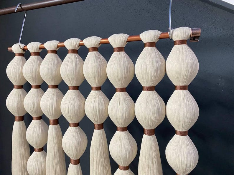 Op de afbeelding: Een witte macram&eacute; wandhanger met bruine lint accenten. De hanger is gemaakt van geknoopt garen en heeft een koperen pijp aan de bovenkant.