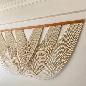 May include: A large wall hanging made of white macrame cord, with a wooden dowel at the top. The macrame is arranged in a series of cascading curves, creating a modern and minimalist design.