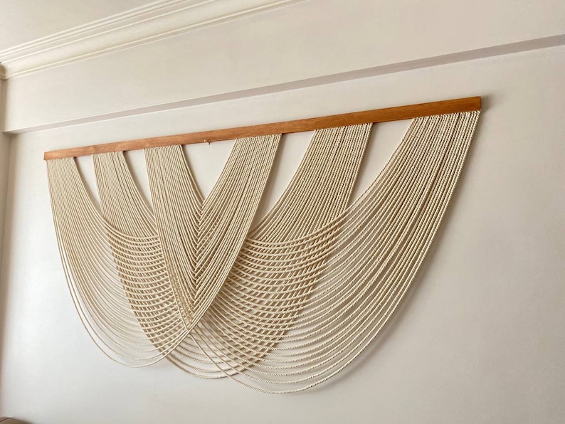 May include: A large wall hanging made of white macrame cord, with a wooden dowel at the top. The macrame is arranged in a series of cascading curves, creating a modern and minimalist design.