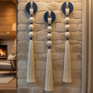 May include: Three decorative wall hangings. Each features a navy blue semi-circle, a series of white spheres, and a long, cream-colored tassel. The hangings are mounted on a textured stone wall, adding a touch of bohemian style to the space.
