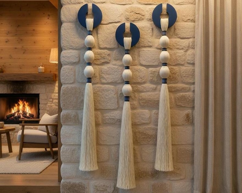 May include: Three decorative wall hangings. Each features a navy blue semi-circle, a series of white spheres, and a long, cream-colored tassel. The hangings are mounted on a textured stone wall, adding a touch of bohemian style to the space.