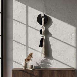 Op de afbeelding: Een wandhanger met een zwart, halvemaanvormig houten bovendeel, een centrale bol verdeeld in een zwarte en witte helft, en een lange kwast bestaande uit afwisselend zwarte en witte vezels, verbonden door kleine zwarte en witte bandjes.