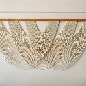 May include: A white macrame wall hanging with a wooden dowel at the top. The macrame is made of a series of knots and loops that create a geometric pattern.