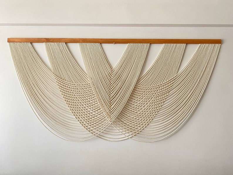 May include: A white macrame wall hanging with a wooden dowel at the top. The macrame is made of a series of knots and loops that create a geometric pattern.