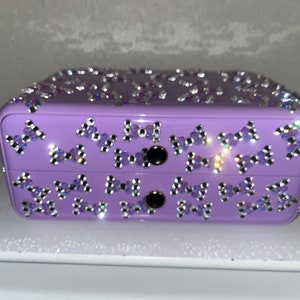 May include: A lavender-colored jewelry box with two drawers. The box is decorated with rhinestones in the shape of bows.