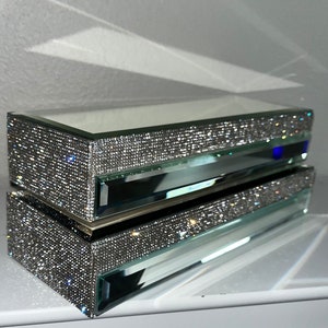 May include: A mirrored jewelry box with a clear glass top and a sparkling rhinestone design. The box has a drawer for storing jewelry.