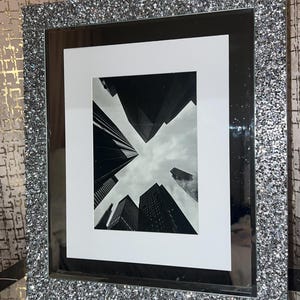 May include: A rectangular picture frame with a sparkling silver glitter border. The frame holds a black and white photograph of tall buildings against a cloudy sky. The frame has a white matting.