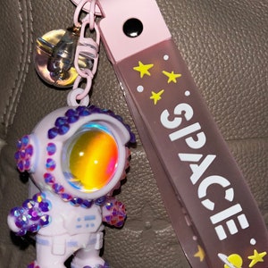 Może przedstawiać: Brelok do kluczy w kształcie astronauty w kolorze różowym i białym z błyszczącym wykończeniem. Brelok ma różową wstążkę z napisem "SPACE" w białych literach. Wstążka ma również żółte gwiazdki i wzór planety.