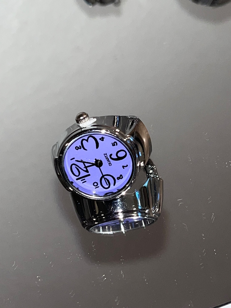 以下が含まれることがあります： 紫色の文字盤と黒い数字が付いた銀色のリングウォッチ。文字盤には「Quartz」と表示され、銀色の秒針が付いています。