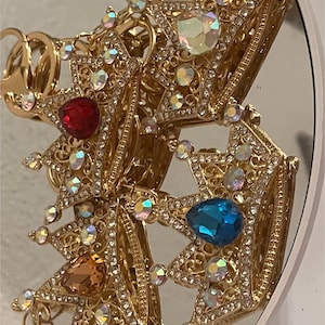 Op de afbeelding: Drie gouden sleutelhangers in de vorm van een kroon met heldere, iriserende strasssteentjes en gekleurde edelsteenaccenten. De sleutelhangers zijn met een metalen ring aan elkaar verbonden.