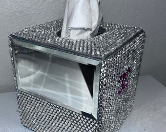 Puede incluir: Un dispensador de pañuelos cuadrado cubierto de brillantes pedrería plateada. Tiene una parte frontal espejada y un logotipo morado. Los pañuelos blancos sobresalen de la abertura superior.