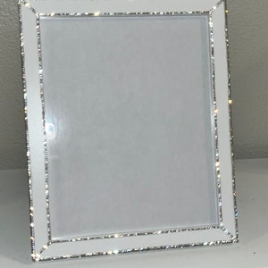 May include: A rectangular white picture frame with a silver glitter border. The frame has a clean, modern design, suitable for displaying photos or artwork. The frame is made of a glossy white material and has a simple, elegant aesthetic.