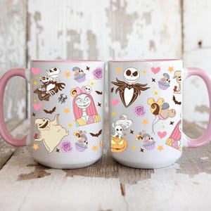 May include: Two white ceramic mugs with pink handles and rims. Each mug features a colorful pattern of characters, cupcakes, bats, and hearts. The mugs are decorated with images of Jack Skellington, Sally, and other characters.