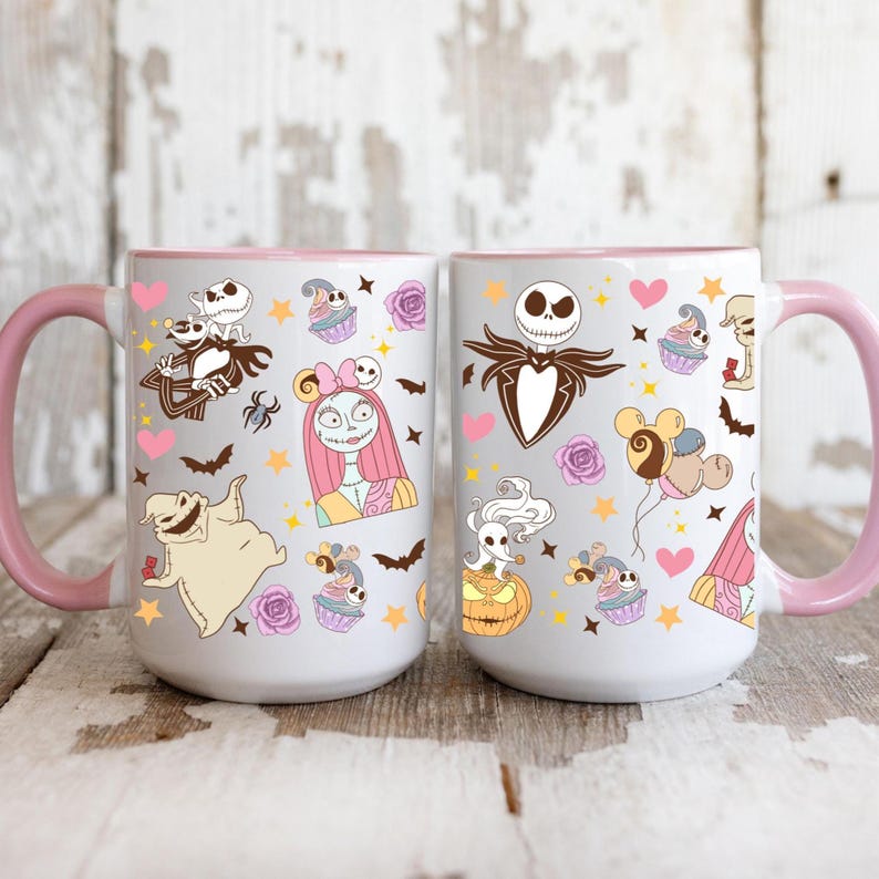 May include: Two white ceramic mugs with pink handles and rims. Each mug features a colorful pattern of characters, cupcakes, bats, and hearts. The mugs are decorated with images of Jack Skellington, Sally, and other characters.