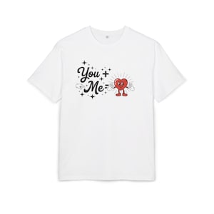 Peut inclure: T-shirt blanc avec un motif graphique représentant le texte "You + Me =" et un personnage de cœur de dessin animé. Le cœur est rouge avec un visage souriant, des bras et des jambes. Le motif comprend des étoiles.