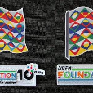 Puede incluir: Dos parches con un diseño de bandera colorida y el texto "UEFA FOUNDATION for children". Un parche incluye el texto "10 YEARS". Las banderas tienen un borde blanco y un patrón de formas rojas, amarillas, azules, verdes y blancas.