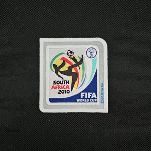 Può includere: Una toppa bianca con un calciatore che calcia un pallone, il testo "South Africa 2010" e il logo della Coppa del Mondo FIFA. La toppa è circondata da un bordo grigio.