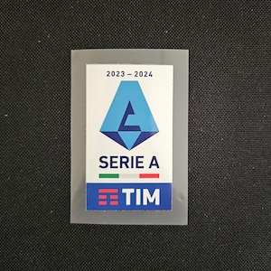 May include: A blue and white patch with the text "2023 - 2024" and "SERIE A" with a logo of a blue triangle with a white letter "A" inside. The patch also has a small Italian flag and the text "TIM" in a white box with three red lines.