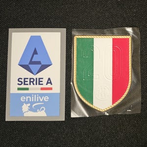 May include: A white sticker with a blue logo and the text "SERIE A" in black. The logo is a stylized letter "A" with a blue and white colour scheme. The sticker also features a small Italian flag and the text "enilive" in black.  A white patch with a green, white, and red Italian flag design. The number "20" is printed in black on the white section of the flag. The patch is surrounded by a gold border.