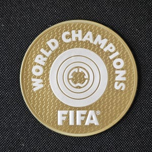 May include: A gold-colored circular patch with the words "WORLD CHAMPIONS" in white, encircling a white emblem. The word "FIFA" is printed in white at the bottom. The patch has a textured surface and is set against a black background.