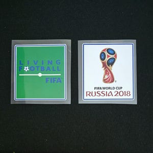 May include: Two iron-on patches for clothing. The left patch is green with the text "Living Football FIFA" in blue. The right patch is white with a red and gold football trophy and the text "FIFA World Cup Russia 2018" in black.